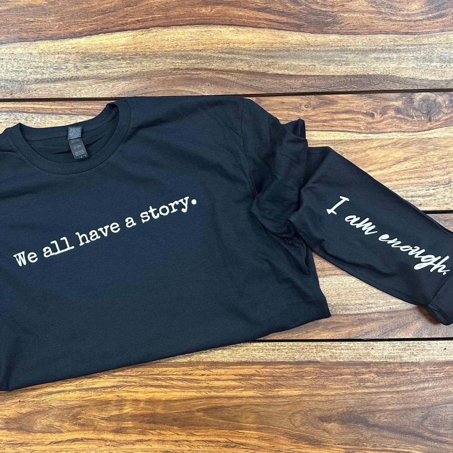Black long sleeve cotton tee.  "We all have a story." is printed across the chest in white ink.  On the left sleeve, printed in cursive font is "I am enough".  Proceeds from the sale of this tee support Call to Freedom, supporting victims of human trafficking.